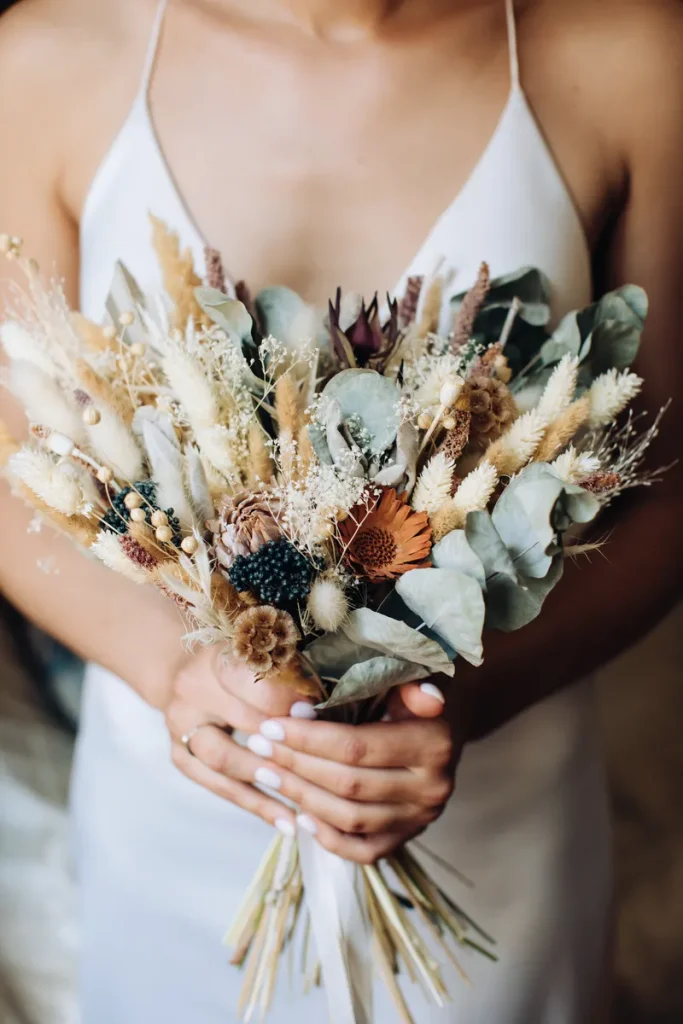 A dried flower wedding bouquet, featuring an arrangement of flowers and some leafy sprigs.